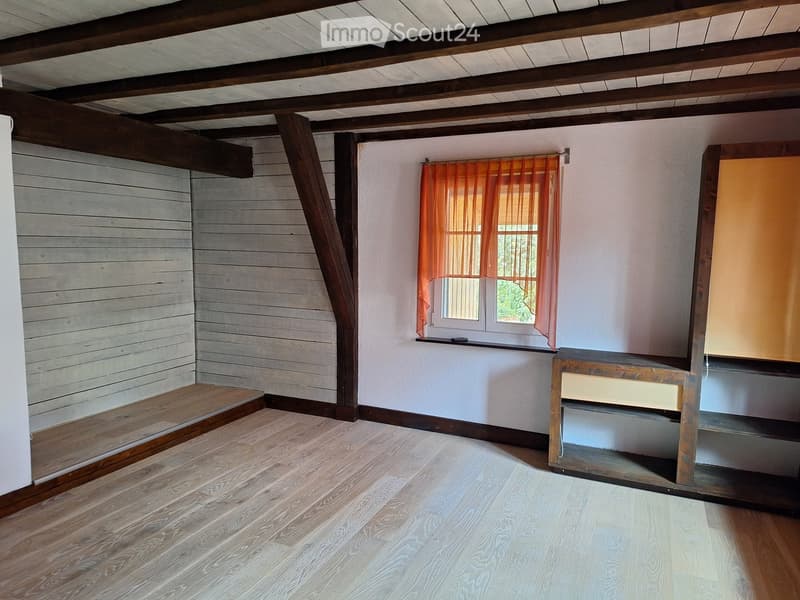 Empty room with wooden flooring, exposed wooden beams on the ceiling, white walls, and a window with orange curtains. There is a small wooden bench or table against the wall.