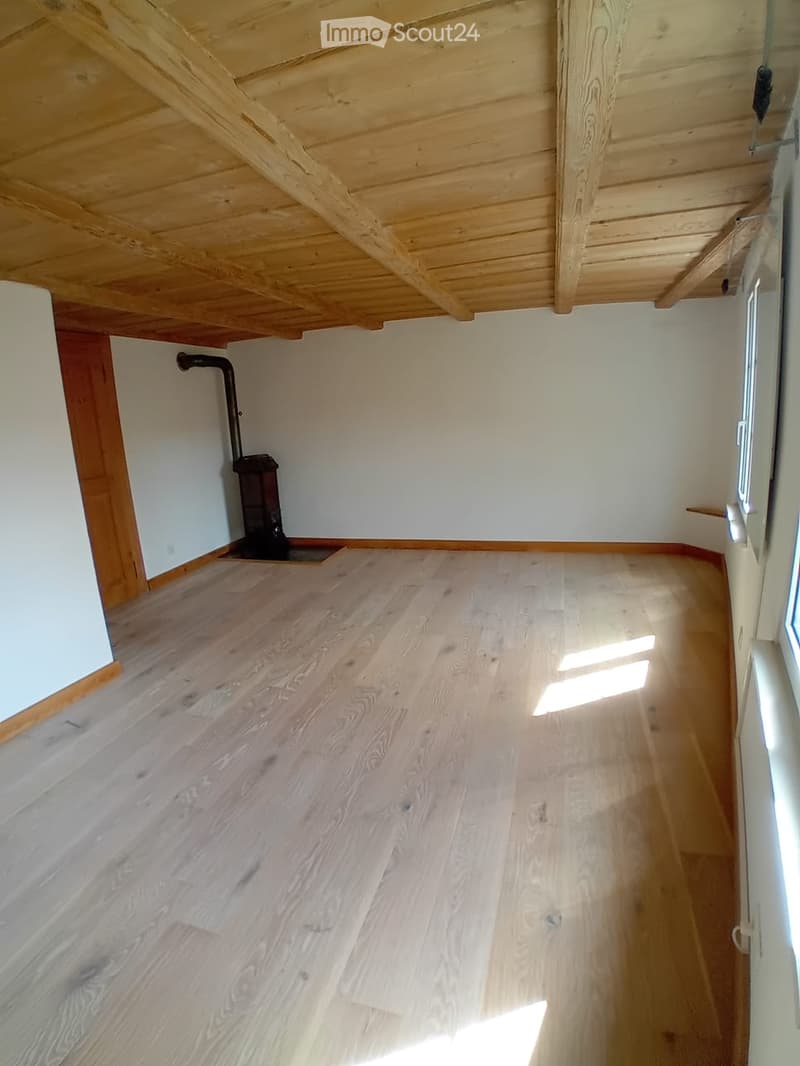 Empty room with wooden flooring and white walls, featuring exposed wooden beams on the ceiling and a small black stove in the corner.