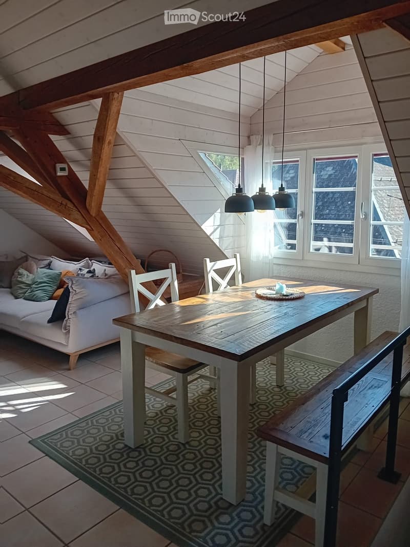 Bright, airy attic space with exposed wooden beams, large windows, a white sofa, a wooden dining table with chairs, and a bench. Natural light floods the room, creating a cozy and inviting atmosphere.