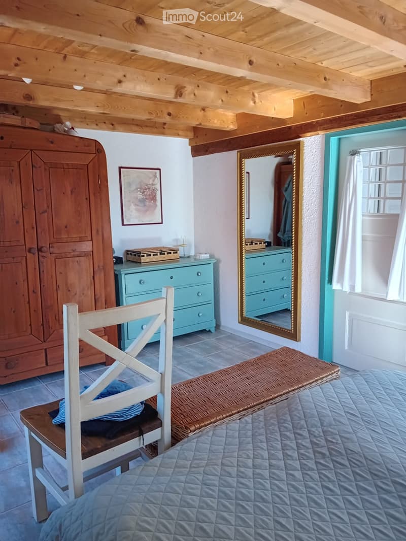 Cozy bedroom with exposed wooden beams, a blue quilted bed, a wooden chair with a cushion, a woven bench, a large mirror, a wooden wardrobe, and a light blue dresser.