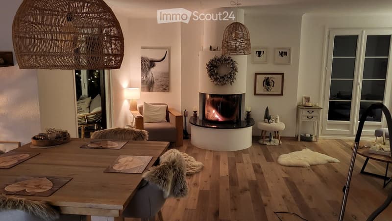 Cozy living and dining area with a central fireplace, wooden dining table, sofa, and decorative elements including a wreath and picture frames.