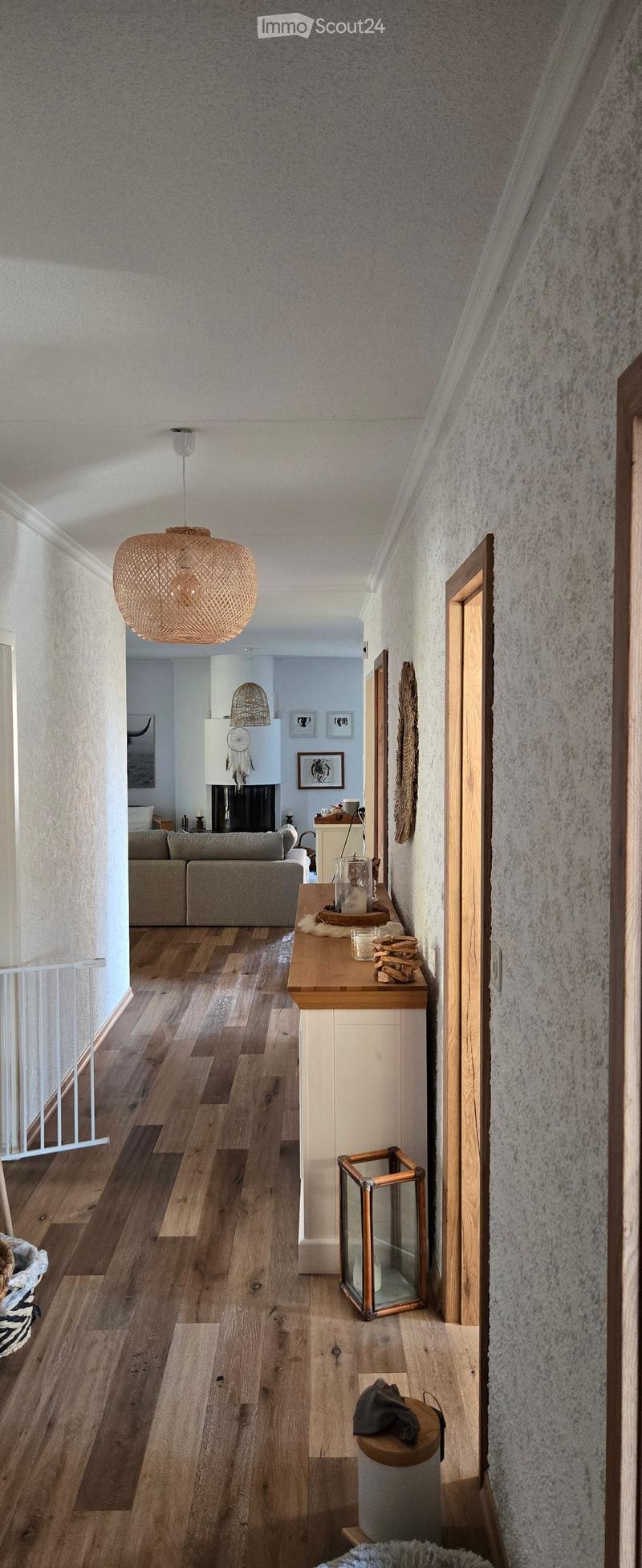 A modern hallway with wooden flooring, white walls, and a hanging woven pendant light. Visible in the background is a living room area with a sofa, fireplace, and decorative items.