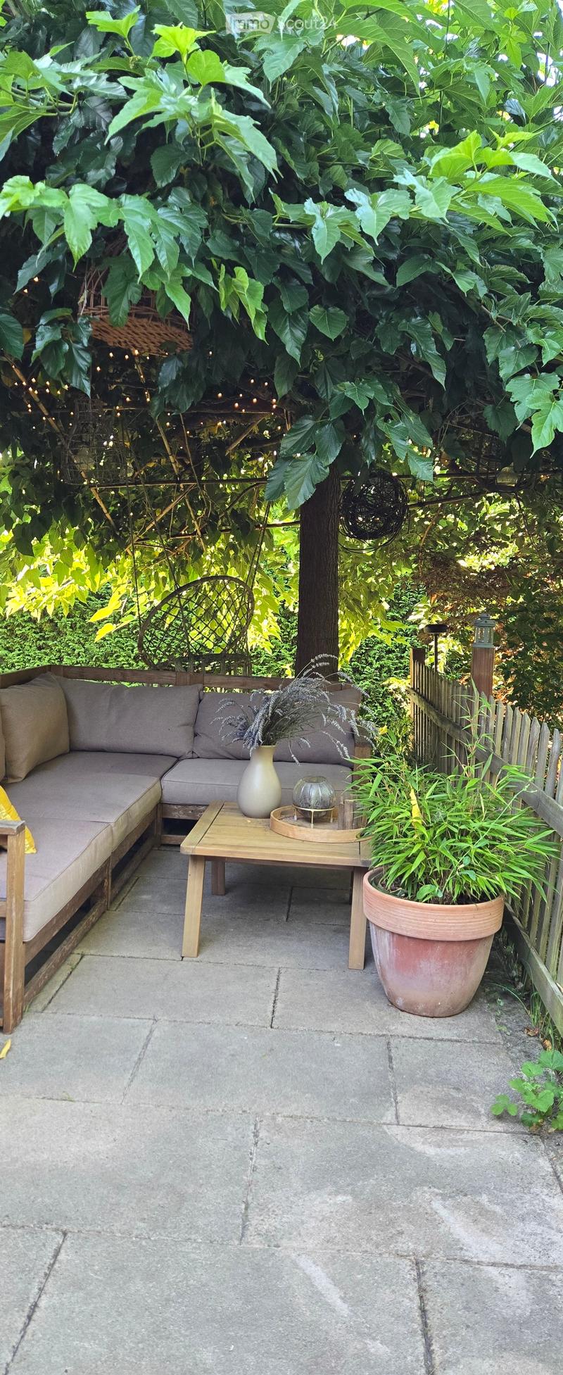 A cozy outdoor seating area with a grey sofa, wooden coffee table, vase with dried flowers, and a large potted plant, surrounded by lush greenery and string lights.