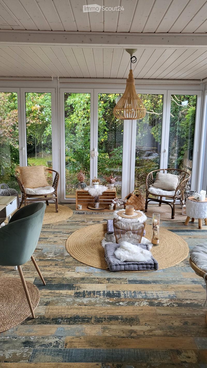 A cozy, well-lit room with large windows overlooking a garden, featuring wooden floors, wicker chairs, a small table, and decorative items.