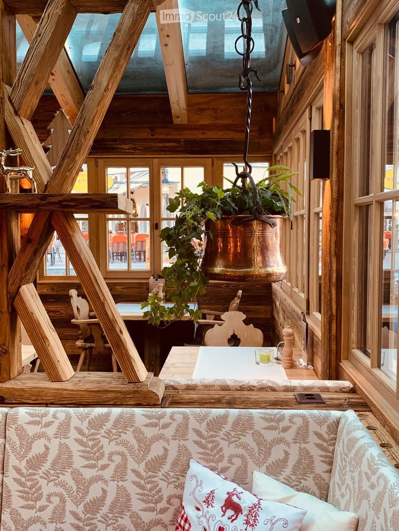 Cozy wooden interior with exposed beams, a patterned sofa, a small table with a candle, and a hanging plant in a copper pot.