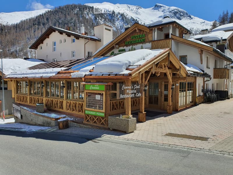 A rustic wooden hotel and restaurant building with snow-covered roofs, large windows, and a sign reading "Pizzeria Restaurant."