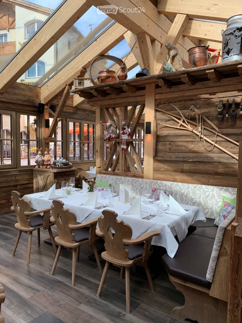 A rustic restaurant interior with wooden beams, a long wooden table set for dining, wooden chairs, a bench, and decorative items including toys and a pot on a shelf.