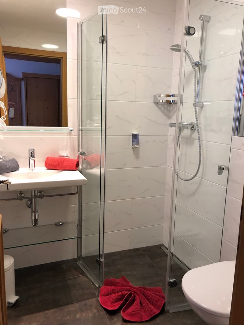 Modern bathroom with white tiles, a glass-enclosed shower, a sink with a mirror, and a bathtub. Red towels are placed on the floor and the sink counter.