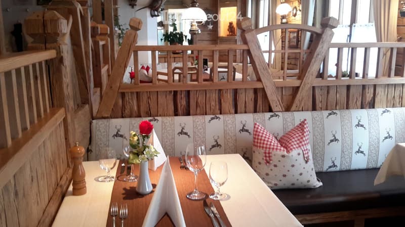 Cozy restaurant setting with wooden tables, wine glasses, napkins, a flower vase, and a bench with deer-patterned cushions and red-and-white checkered pillows.
