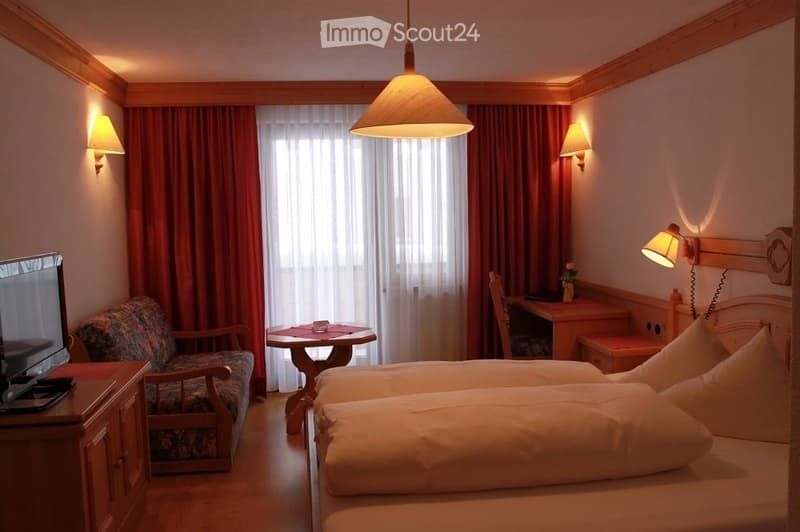 Cozy bedroom with wooden furniture, two single beds, a small table, a chair, a desk, and a TV screen. The room features warm lighting from wall-mounted lamps and a central pendant light, with red curtains covering the window.