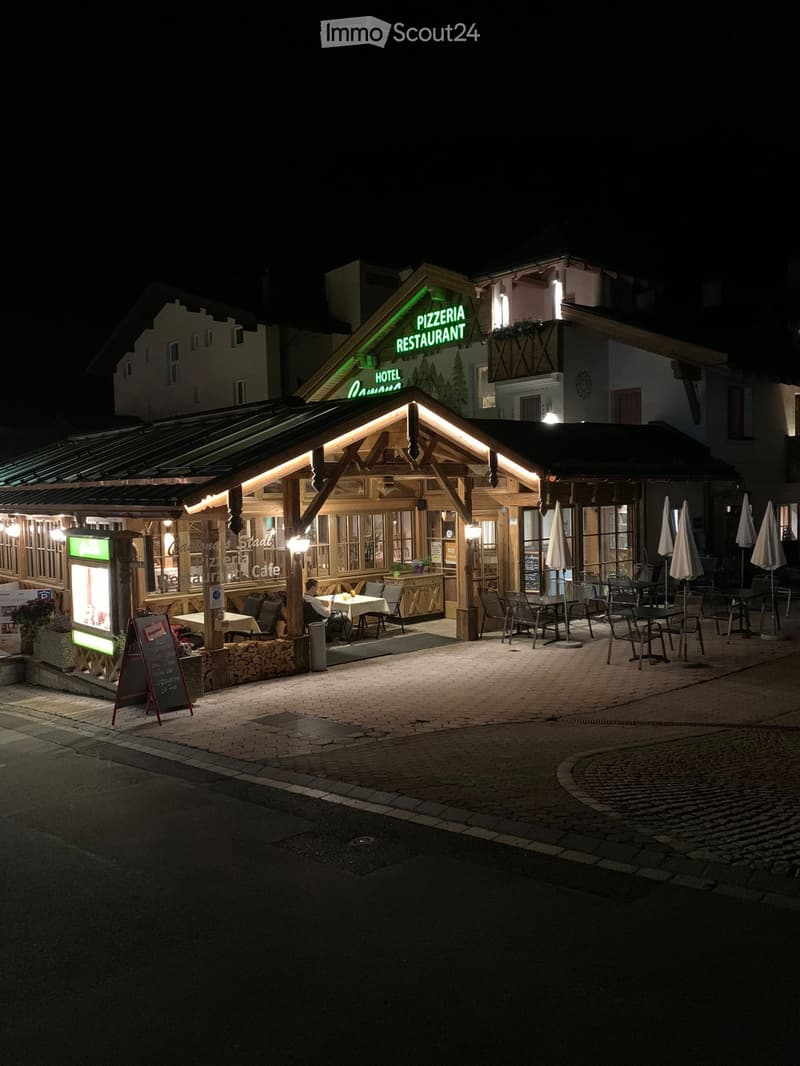 Nighttime view of a restaurant with a covered outdoor seating area, illuminated signage reading "Pizzeria Restaurant," tables, chairs, and umbrellas.