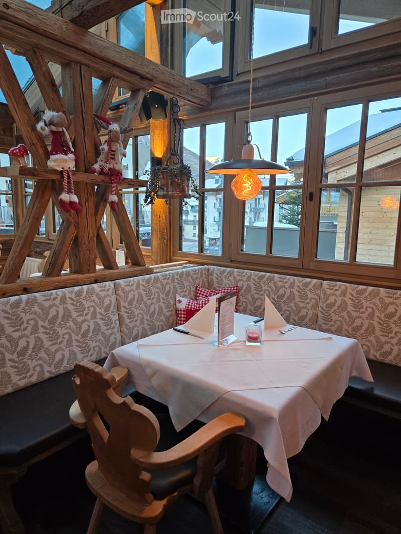 Cozy restaurant interior with wooden beams, large windows, patterned sofa seating, and a neatly set dining table with menu and candle.