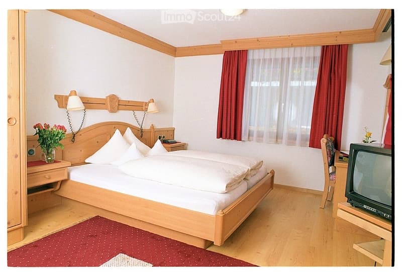 Cozy bedroom with a wooden bed, nightstand, lamp, chair, television, and a vase with flowers on the nightstand.