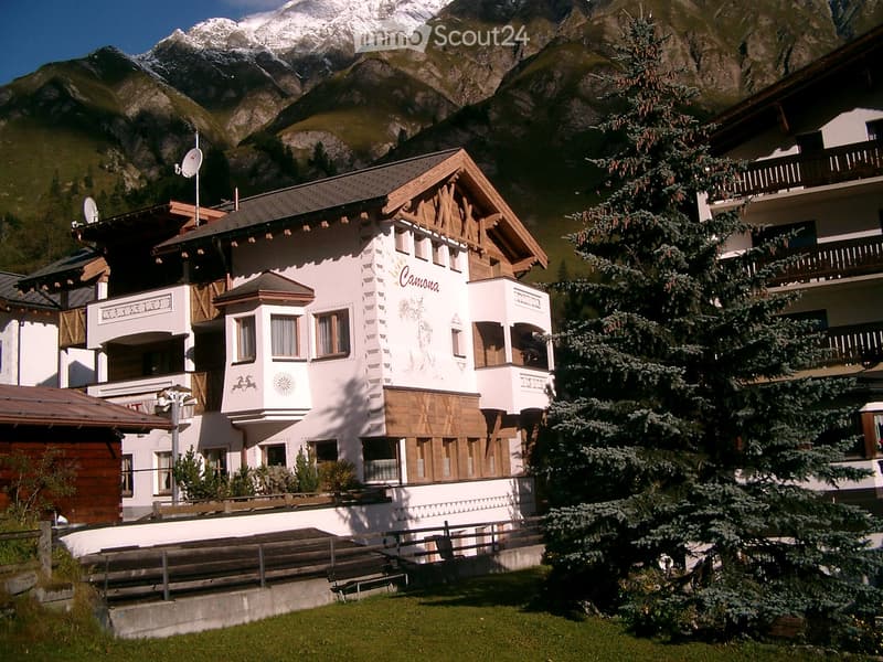 A multi-story building with a white facade and wooden accents, featuring balconies and a prominent sign reading "Camona" against a backdrop of snow-capped mountains.