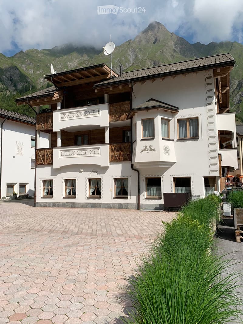 A large, multi-story white house with wooden balconies and intricate decorative elements, set against a backdrop of mountains.