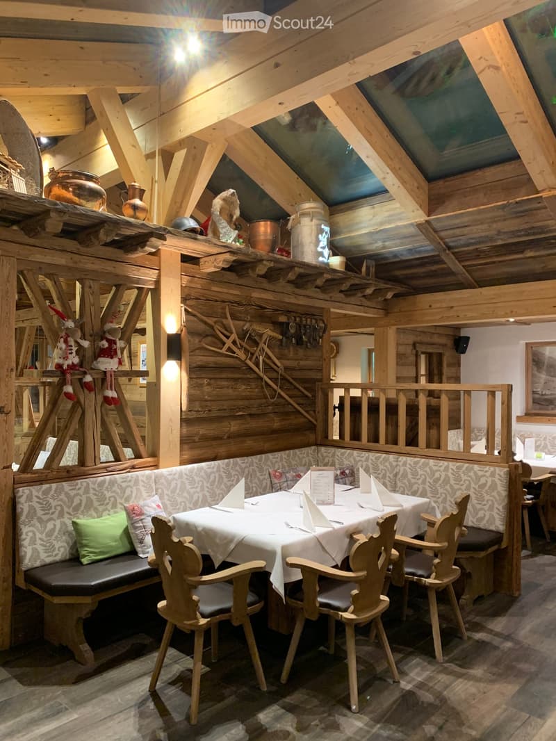 Cozy restaurant with wooden beams, stone floor, and rustic decor. Features a long wooden table with chairs, a bench seating area, and decorative vases on shelves.