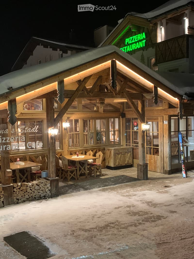 Wooden outdoor seating area of a restaurant with tables, chairs, and a stack of firewood, surrounded by wooden structures and snow-covered ground.