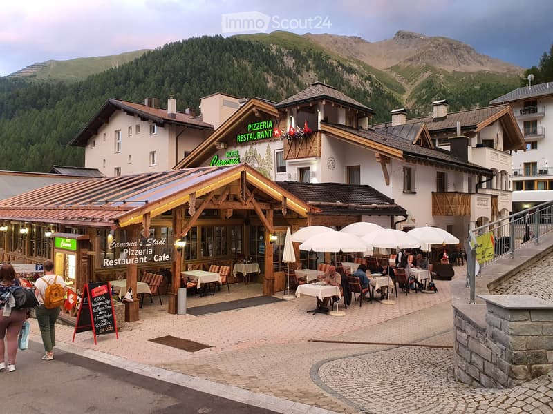 Outdoor seating area of a restaurant with tables, chairs, and umbrellas, surrounded by wooden structures and a scenic mountain backdrop.