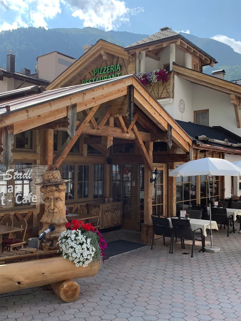 Outdoor seating area of a restaurant with wooden tables, black chairs, and a large umbrella, surrounded by flowers and a scenic mountain backdrop.