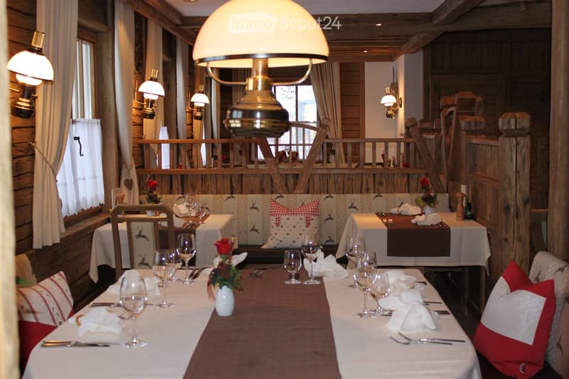Cozy restaurant interior with wooden walls, large windows, and a long dining table set with wine glasses, napkins, and floral arrangements. A sofa with pillows is visible in the background.