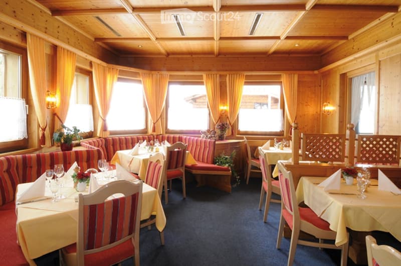 A cozy restaurant interior with wooden walls and ceiling, large windows with curtains, tables set with tablecloths, napkins, wine glasses, and cutlery, and red and white striped sofas along the walls.