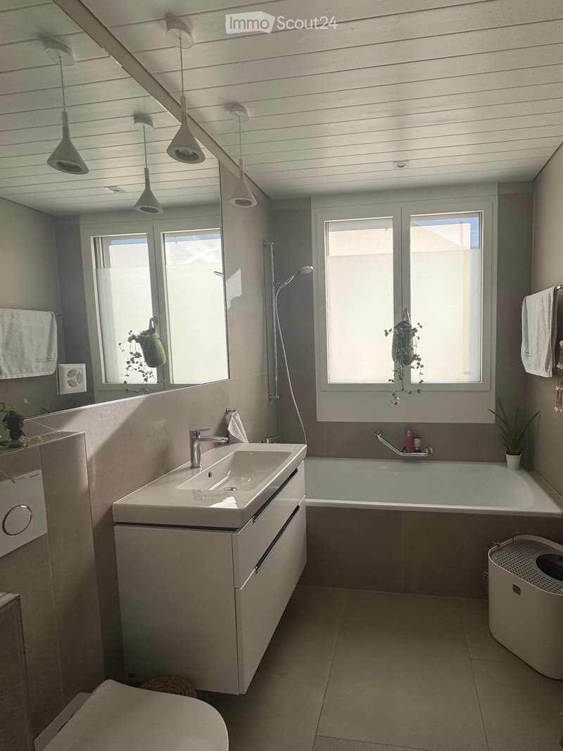 Modern bathroom with a white sink, bathtub, toilet, and large windows. Features a sleek faucet, modern light fixtures, and a mirror above the sink.