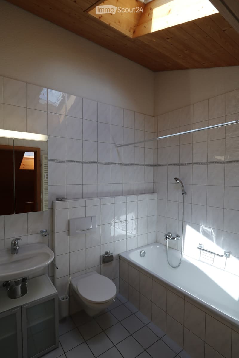 Modern bathroom with white tiles, a bathtub, toilet, sink, and a large mirror. The room features a skylight and a wooden slanted ceiling.