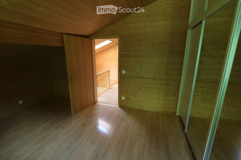 Empty wooden hallway with polished floor, wooden walls, and a door leading to another room. Natural light streams in from an open doorway at the end.