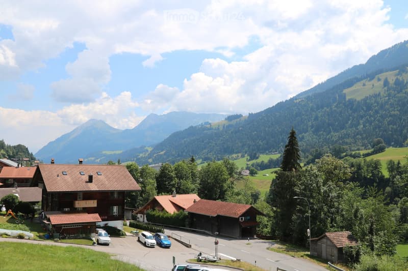 A cluster of traditional wooden houses with red-tiled roofs in a picturesque alpine village, surrounded by lush green trees and mountains.