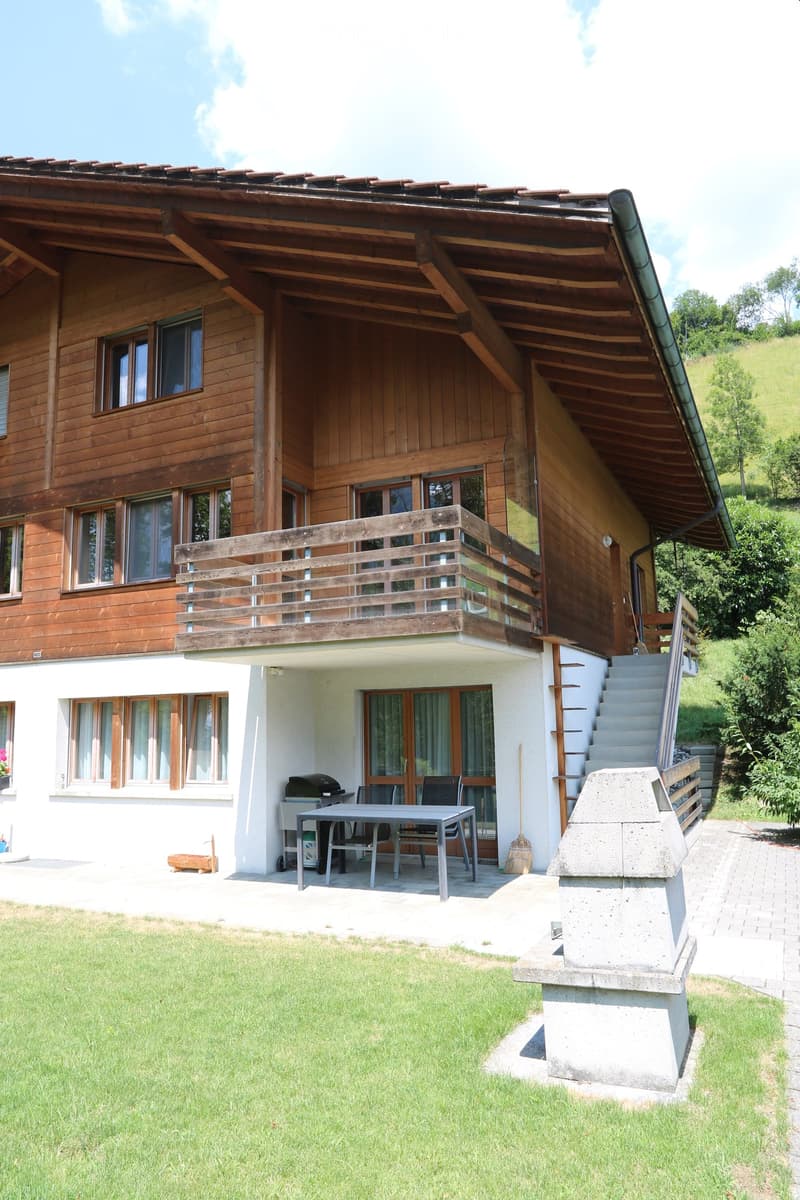 A well-maintained garden area with a wooden house in the background, featuring a patio with a table, chairs, and a grill, along with a staircase leading to an upper balcony.