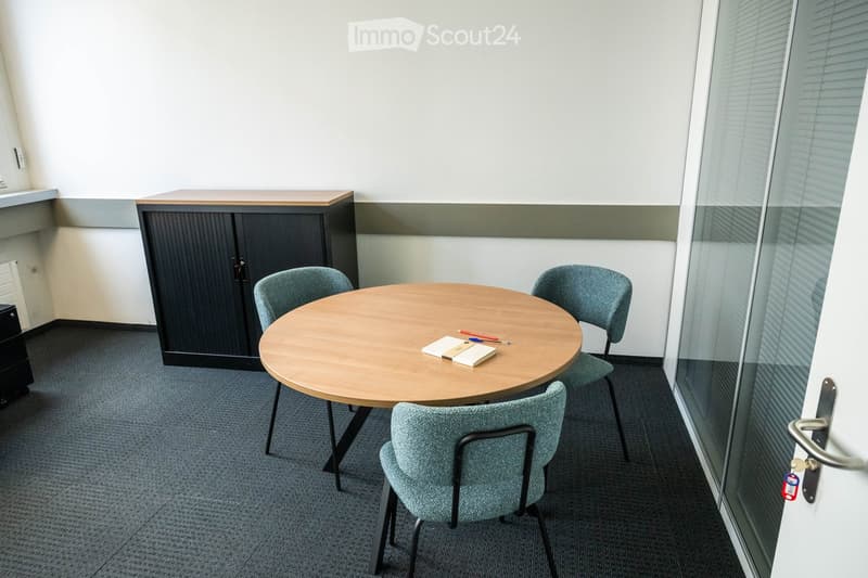 A small, modern meeting or break room with a round wooden table, four grey chairs, a dark wooden cabinet, and a glass wall with a door.