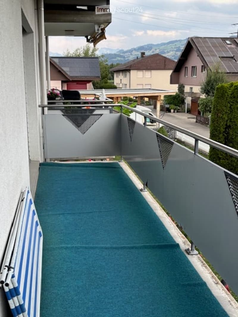 Balcony with green carpeting, metal railing, and a view of the surrounding houses and mountains.