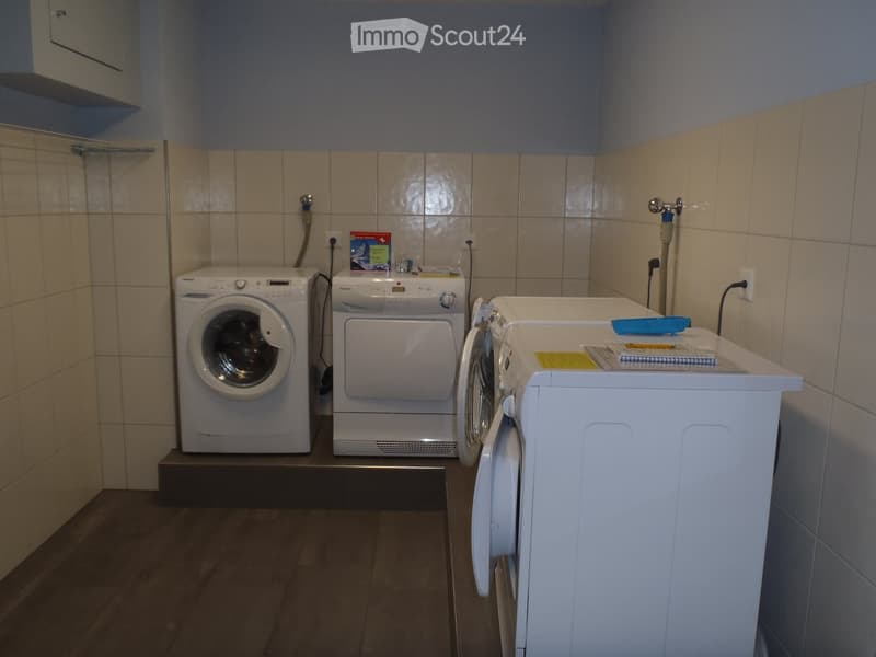 A small bathroom with a washing machine, dryer, and sink. The walls are tiled, and there is a book on the sink counter.