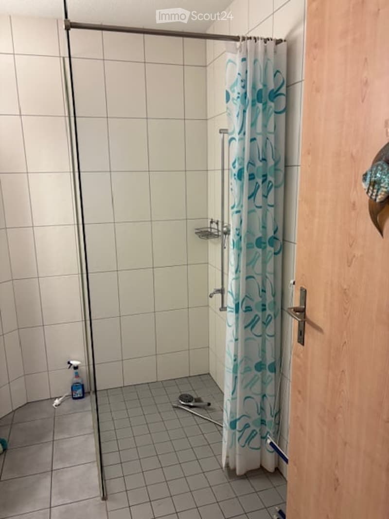 Shower area with white tiled walls and floor, glass shower enclosure, blue and white shower curtain, shower rack, cleaning supplies on floor.