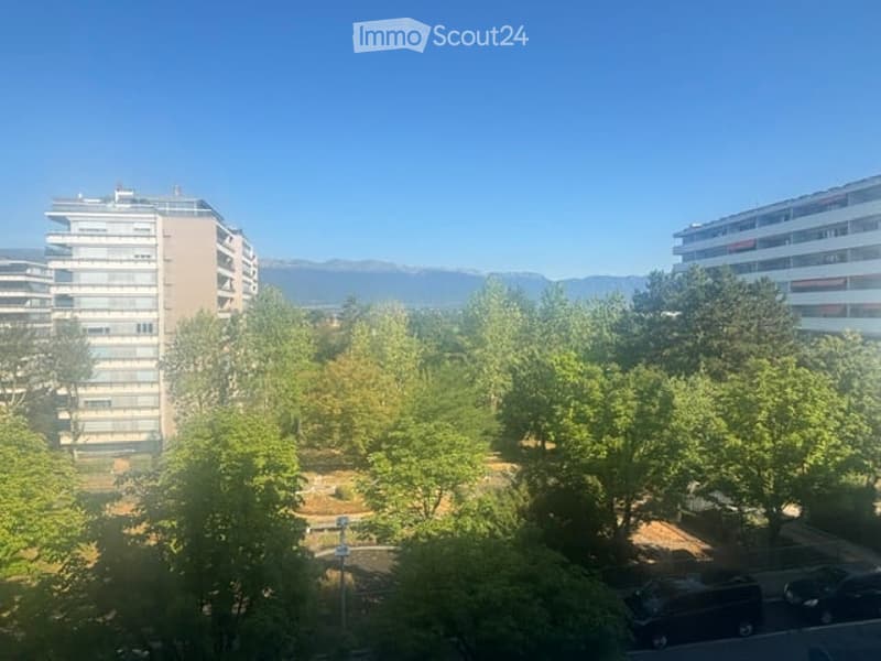 Balcony view overlooking a park with trees, buildings, and mountains in the background.
