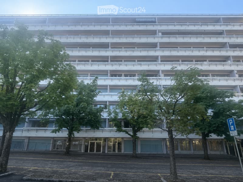 A multi-story residential building with white facade, balconies, and trees in front.