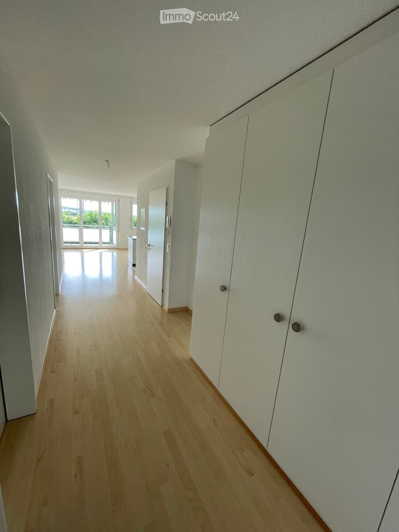 Empty hallway with light wooden flooring, white walls, and built-in closet doors. Natural light streams in from a large window at the end of the corridor.