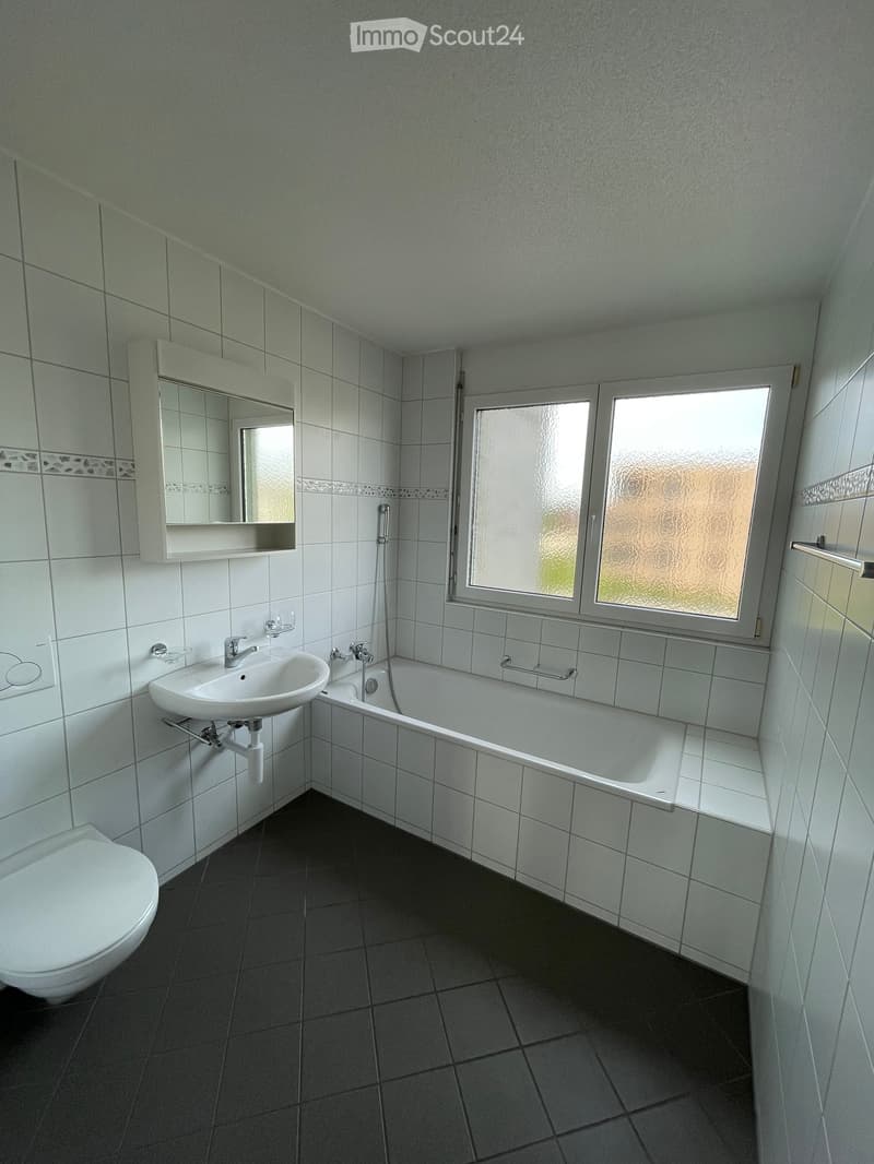 Modern bathroom with white tiles, a toilet, a sink, a bathtub, and a mirror above the sink. The room has a window with frosted glass and a dark tiled floor.