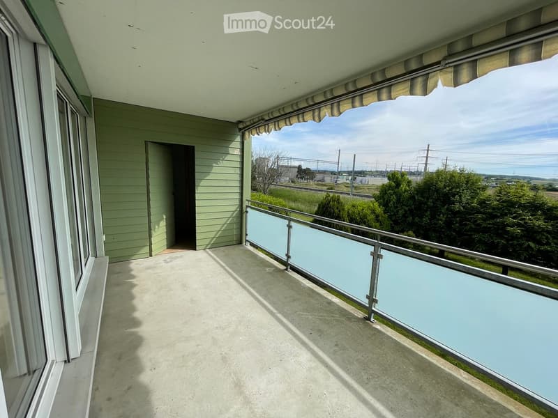 Balcony with a modern glass railing, concrete floor, and a retractable awning. Offers a view of a green landscape and distant railway tracks.