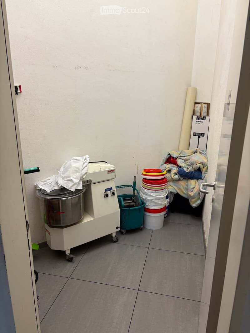 A small utility room with a large industrial mixer, several stacked buckets, a mop bucket, and a folded blanket on the floor.