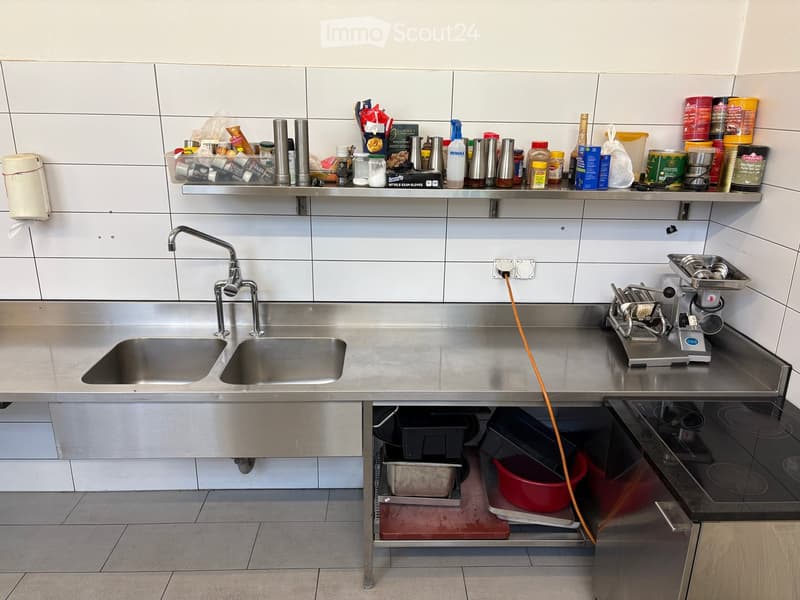 Stainless steel kitchen with double sink, faucet, shelves stocked with various items, coffee machine, and oven.