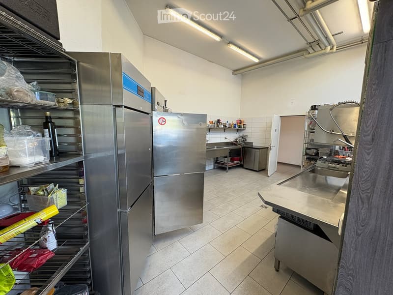 Commercial kitchen with stainless steel appliances, including a large refrigerator, oven, sink, and counter. Shelves hold various supplies and ingredients.