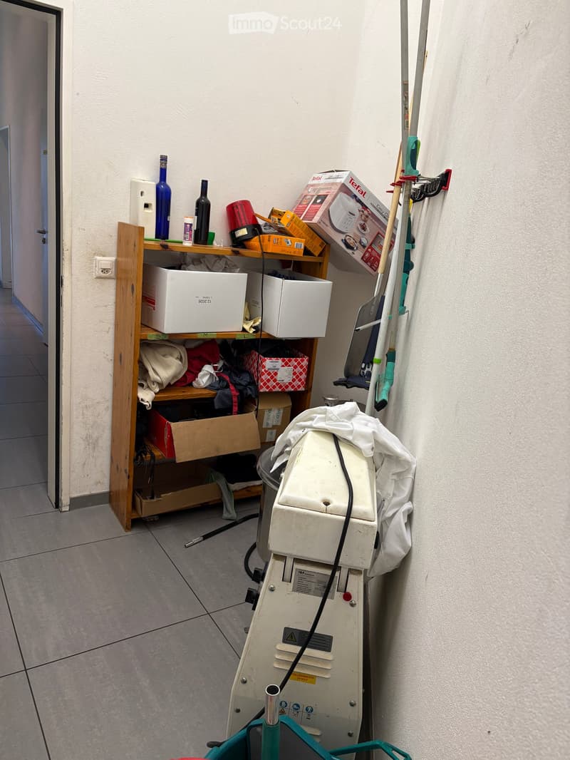 A small utility or storage room with a wooden shelf holding various items, including bottles, boxes, and cleaning supplies. A large cleaning machine is positioned on the floor, and cleaning tools are mounted on the wall.