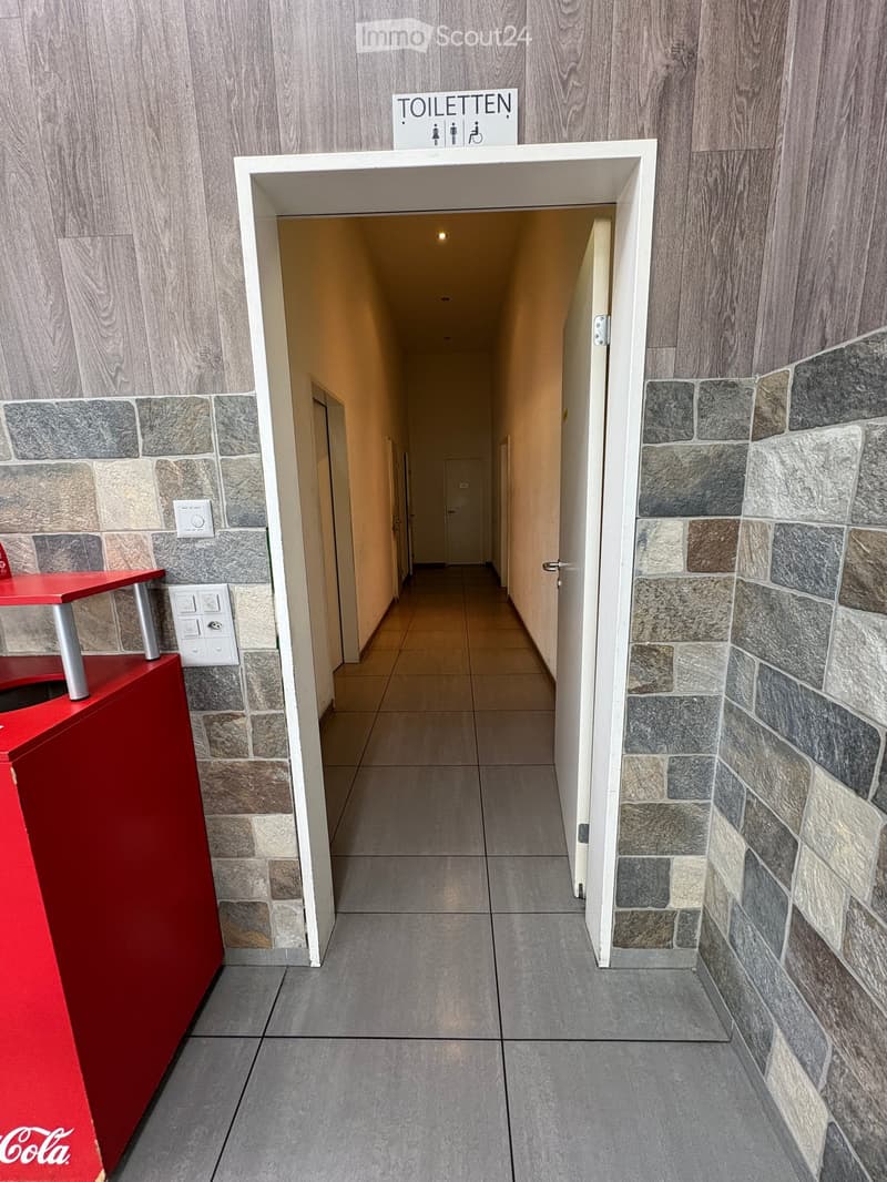 Clean, modern hallway with tiled floors, stone-patterned walls, and a red Coca-Cola stand on the left side. A sign above a doorway reads 'TOILETTEN' with symbols for male and female restrooms.