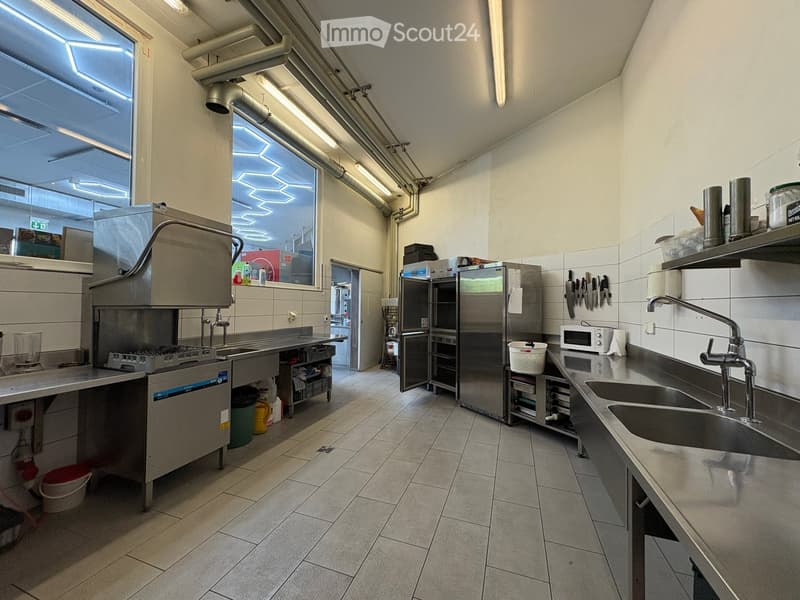 Commercial kitchen with stainless steel appliances, including a sink, refrigerator, microwave, and oven. The space features tiled floors and walls, with various kitchen tools and utensils visible.