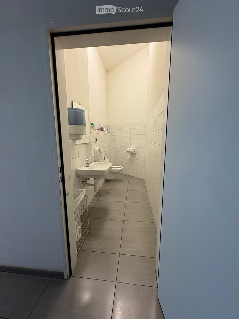 Clean, modern bathroom with white tiled walls and floor, featuring a sink, toilet, paper towel dispenser, and hand dryer. A small shelf holds toiletries.