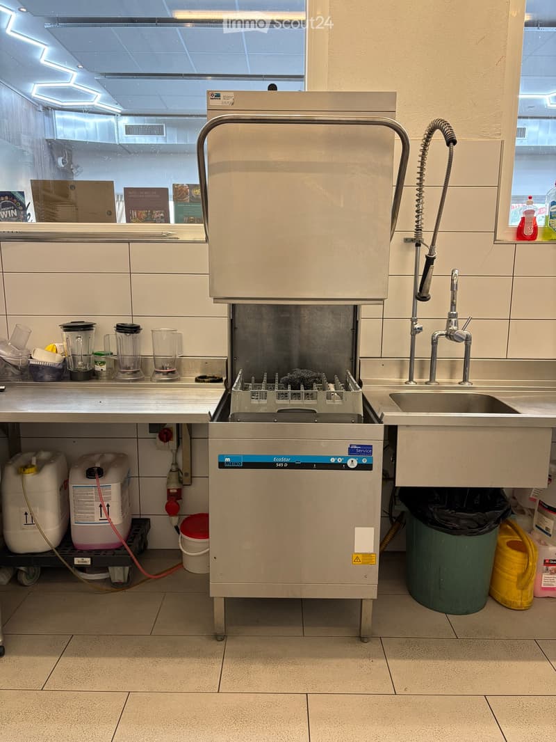 Commercial kitchen with stainless steel dishwasher, sink, blender, and various cleaning supplies.