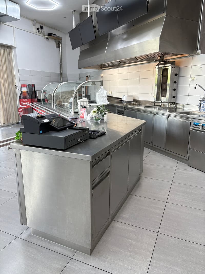 Modern commercial kitchen with stainless steel appliances, a central island, and a display case. Includes a cash register, sink, and various kitchen equipment.