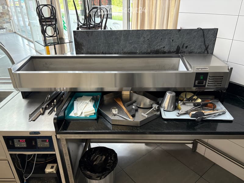 Stainless steel kitchen counter with sink, hood, and various utensils including trays, spoons, and a wooden spoon. A trash bag is placed on the floor.