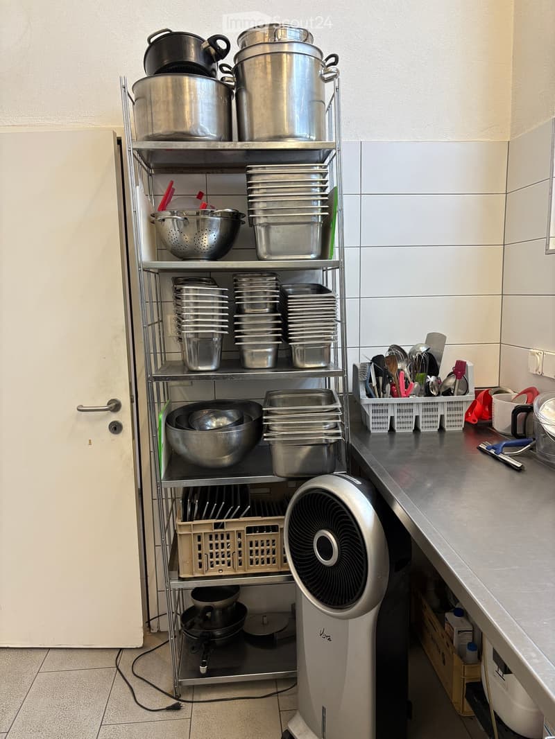 A kitchen with a metal shelving unit filled with pots, pans, and bowls. A fan is placed on the floor, and a counter holds various kitchen utensils and a sink.